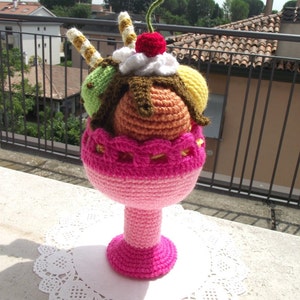 May include: A crocheted ice cream sundae with a pink bowl, brown chocolate sauce, green, orange, and yellow scoops of ice cream, a cherry on top, and two yellow and brown candy sticks.