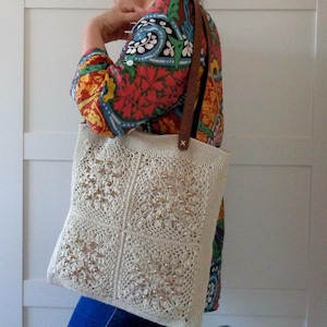 Peut inclure: Sac fourre-tout en crochet crème avec un motif carré et de petites perles décoratives. Le sac est doté de lanières en cuir marron. Une personne porte le sac et porte une chemise à motifs colorés.
