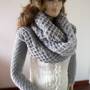 May include: A gray knitted cowl neck scarf with a waffle stitch pattern. The scarf is worn by a model with long brown hair.