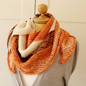 May include: A triangular knitted shawl in shades of orange, white, and brown. The shawl is draped over a mannequin and has a striped pattern.