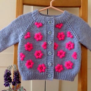 May include: A pale blue knitted cardigan adorned with vibrant pink flower appliqués. The baby's sweater features long sleeves, a round neckline, and is fastened with light blue buttons. The floral design adds a charming touch to this cosy garment.
