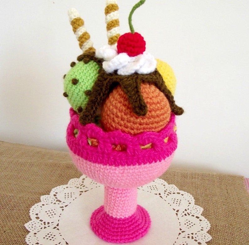 May include: A crocheted ice cream sundae with three scoops of ice cream in a pink crocheted bowl. The ice cream scoops are green, orange, and yellow. The sundae is topped with a white crocheted dollop of whipped cream, a red crocheted cherry, and two brown and white crocheted sticks.