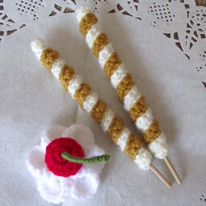 May include: Two crocheted sticks with white and yellow stripes, and a white crocheted flower with a red center. The sticks have wooden dowels attached to the ends.