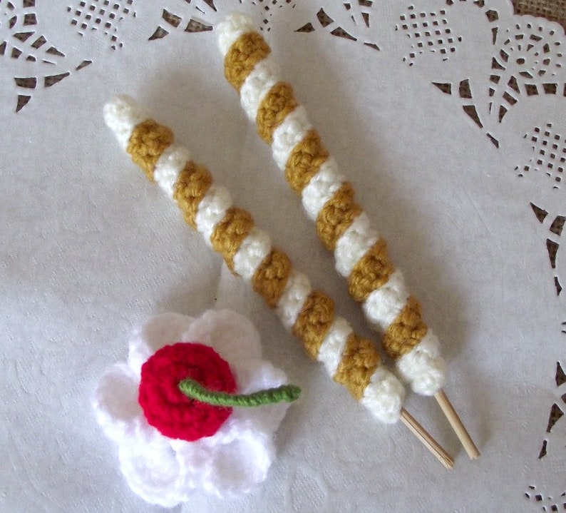 May include: Two crocheted sticks with white and yellow stripes, and a white crocheted flower with a red center. The sticks have wooden dowels attached to the ends.