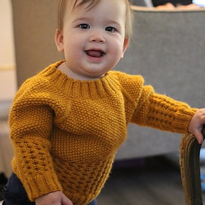 May include: A smiling baby wearing a yellow knitted sweater with a textured pattern and blue pants.