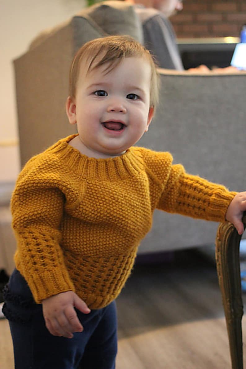 May include: A smiling baby wearing a yellow knitted sweater with a textured pattern and blue pants.