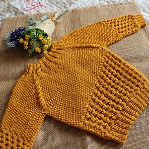 May include: A mustard yellow knitted sweater with a textured stitch pattern. The sweater has long sleeves and a round neckline.
