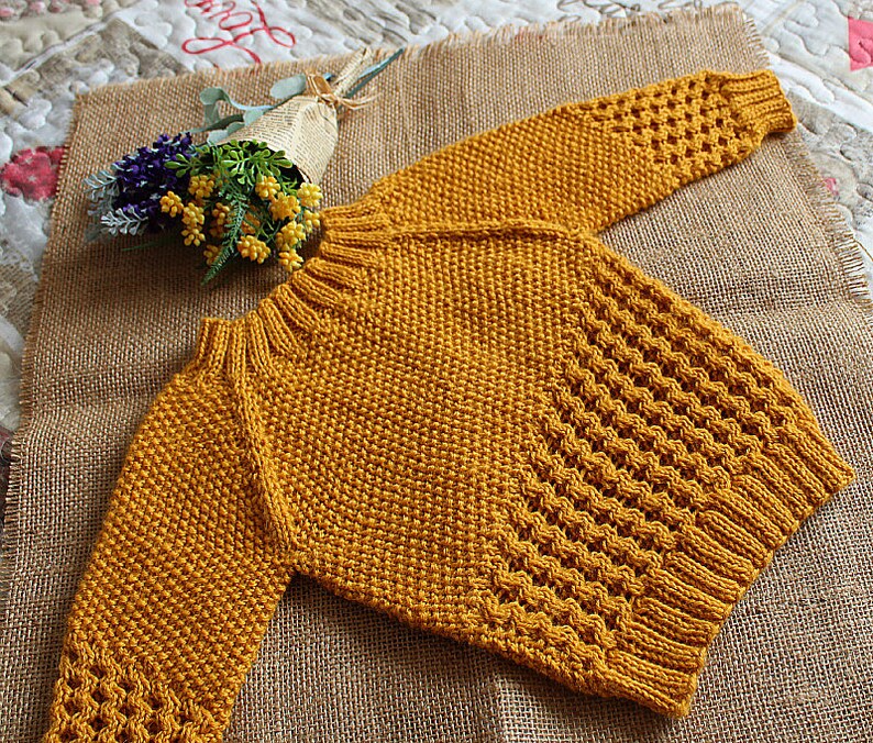 May include: A mustard yellow knitted sweater with a textured stitch pattern. The sweater has long sleeves and a round neckline.