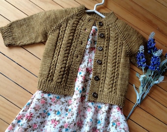 May include: A brown knitted cardigan with a cable knit pattern and button closure. The cardigan is worn over a white dress with a floral print.