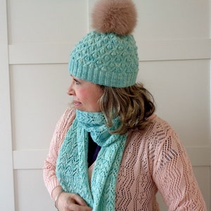 May include: A teal blue knitted hat with a fluffy brown pom pom on top. The hat is paired with a matching teal blue knitted scarf.