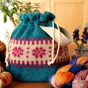 May include: Teal knitted drawstring bag with a white strap and a pink and cream snowflake pattern. The bag is filled with colorful yarn.