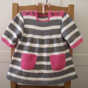 Puede incluir: Un vestido de punto a rayas grises y blancas con ribetes y bolsillos rosas. El vestido tiene un escote redondo y mangas cortas.