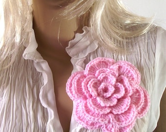 Puede incluir: Un broche de flor rosa tejido a crochet, usado en una blusa blanca con volantes.