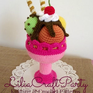 May include: A crocheted ice cream sundae with a pink bowl, three scoops of ice cream, a cherry on top, and two chocolate-covered waffle cones.