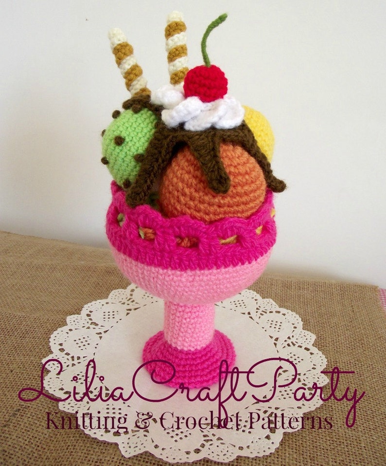 May include: A crocheted ice cream sundae with a pink bowl, three scoops of ice cream, a cherry on top, and two chocolate-covered waffle cones.