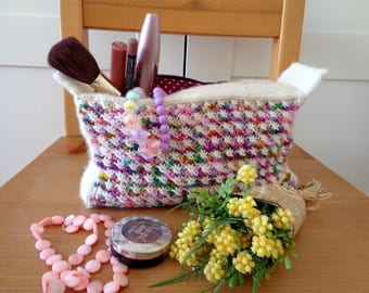 May include: A white knitted zippered pouch with a colorful, speckled pattern. The pouch is filled with makeup brushes, lipstick, and other cosmetics. A bouquet of yellow flowers sits in front of the pouch.