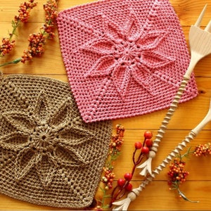 Può includere: Due strofinacci all'uncinetto, uno marrone e uno rosa, con un motivo a stella. Gli strofinacci sono su una superficie di legno con utensili di legno.