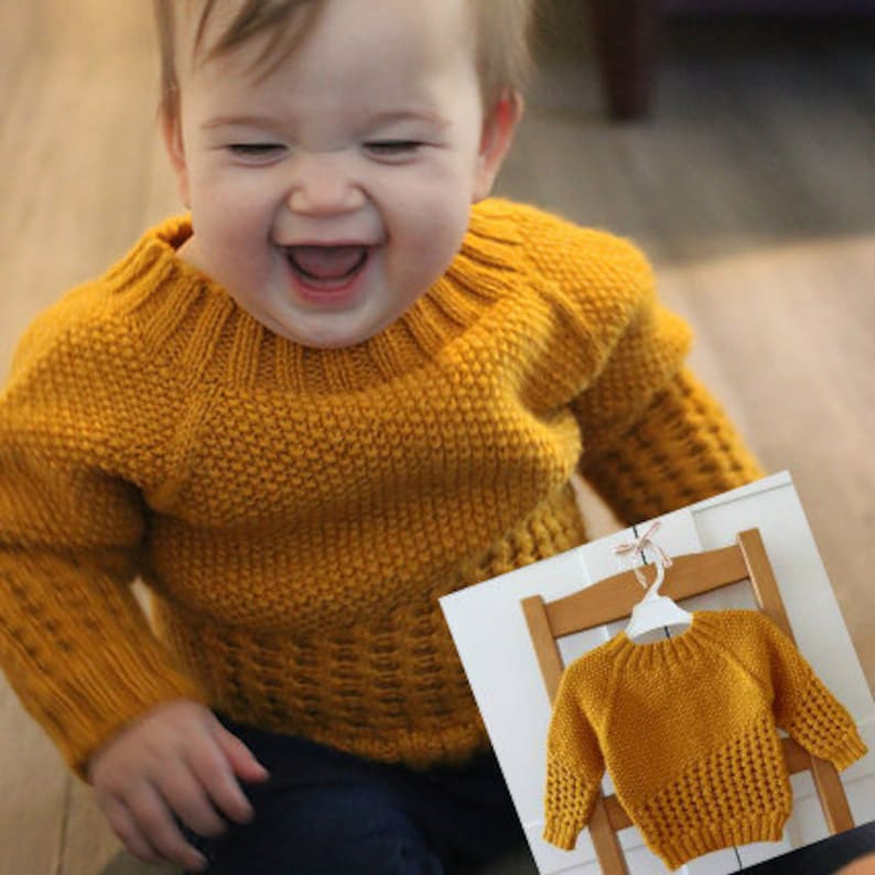 May include: A smiling child wearing a yellow knitted sweater with a textured pattern. The sweater has a round neckline and long sleeves.