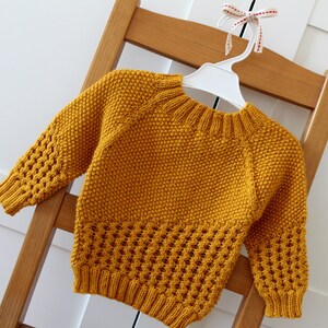 May include: A mustard yellow knitted sweater with a round neckline and long sleeves. The sweater has a textured pattern and a lace-like design at the bottom.