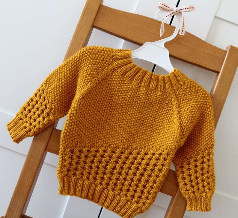 May include: A mustard yellow knitted sweater with a round neckline and long sleeves. The sweater has a textured pattern and a lace-like design at the bottom.