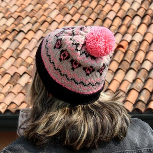May include: A pink and black knit beanie with a pom pom on top. The beanie has a geometric pattern with pink and black accents.