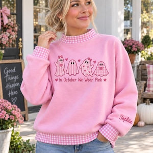 Op de afbeelding: Een lichtroze sweatshirt met een opstaande kraag en boorden in een roze-wit vichy-ruitpatroon. De voorzijde toont vier spookillustraties en de tekst "In October We Wear Pink". De naam "Sophia" is geborduurd op de linkermouw.