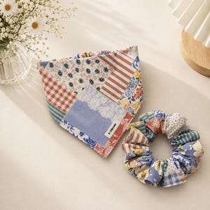 May include: A triangular pet bandana and a matching fabric scrunchie featuring a patchwork design of blue, white, and orange floral, striped, and gingham patterns. The bandana includes a small white fabric tag with the text D Bloom printed in black.