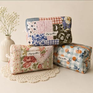 May include: Three rectangular quilted pouches with zippered closures. One features a patchwork of floral and gingham patterns, another displays a rose floral print, and the third shows an orange fruit pattern on a light blue gingham background.