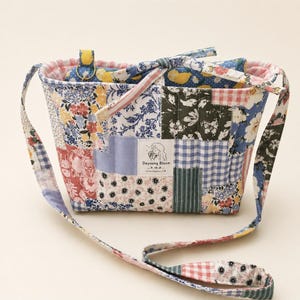 May include: A quilted patchwork shoulder bag featuring a variety of floral, gingham, and striped fabric patterns in blue, pink, yellow, and green. A white rectangular label on the front displays the text Dayoung Bloom with a line drawing.
