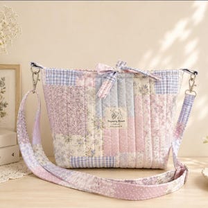 May include: A quilted crossbody bag featuring a patchwork design of light purple, pink, and blue floral and gingham patterns. The bag has a fabric bow at the top center, a matching adjustable shoulder strap, and a rectangular label reading Dayoung Bloom.