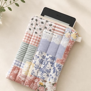May include: A quilted fabric sleeve for an e-reader featuring a patchwork design of floral, gingham, and striped patterns in muted pink, blue, and white tones. A white label with the text Dayeong Bloom is stitched on the front with a gold zipper.