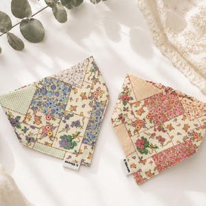 May include: Two triangular pet bandanas featuring a patchwork design of floral patterns. The left bandana displays blue, yellow, and green flowers on a cream background. The right bandana features pink, orange, and yellow flowers. Both have a D Bloom tag.