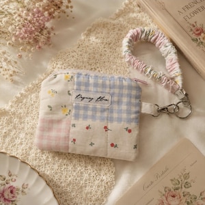 May include: A quilted fabric pouch featuring a patchwork design of light blue gingham, pink gingham, and floral patterns with yellow and red flowers. A label reads Dayning Bloom. A matching fabric wrist strap is attached with a silver metal clasp.