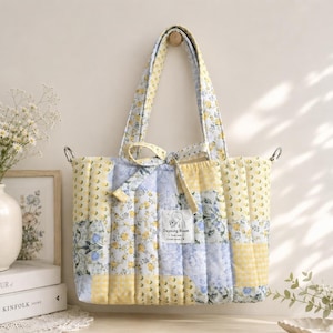 May include: A quilted tote bag featuring a patchwork design of yellow and light blue floral patterns. The bag has two shoulder straps, a tie closure at the top, and a white rectangular fabric label with the text Dayoung Bloom hand made.