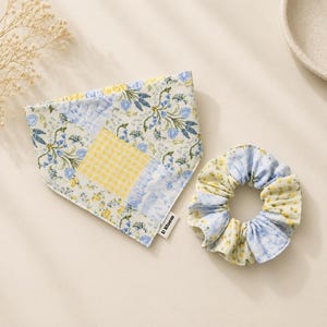 May include: A triangular pet bandana and a matching fabric scrunchie featuring a patchwork design of blue floral patterns, yellow gingham checks, and small yellow flowers. A small white tag with the text D Bloom is attached to the bandana.