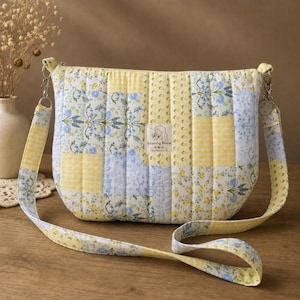 May include: A quilted crossbody bag featuring a patchwork design of yellow gingham, blue floral patterns, and yellow flower prints. The bag has a rectangular label in the center with the text Dayoung Bloom and a long, matching fabric shoulder strap.