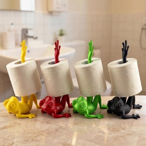 Op de afbeelding: Vier eigenzinnige toiletrolhouders in geel, rood, groen en zwart. Elke houder heeft een kikkerachtige basis met uitgestrekte poten en een hand die een rol wit toiletpapier vasthoudt. Op een lichtgekleurd oppervlak.