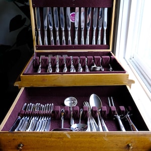 May include: A wooden silverware chest with two drawers, open to reveal a complete set of polished silverware. The interior is lined with a deep purple velvet, and the silverware is neatly arranged in designated slots. The chest is made of light-colored wood.