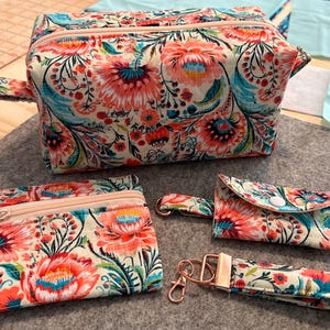 May include: A floral print cosmetic bag, a small zippered pouch, a small wallet, and a wristlet with a gold-tone clasp. The items are on a gray felt surface. The floral pattern includes red, pink, and teal flowers.