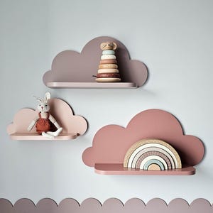 Peut inclure: Trois étagères en forme de nuage, dans des tons de rose et de mauve, sont fixées sur un mur bleu clair. Elles présentent un jouet à empiler en bois, un lapin en peluche et un arc-en-ciel en bois. Ces étagères offrent un rangement décoratif et fonctionnel.