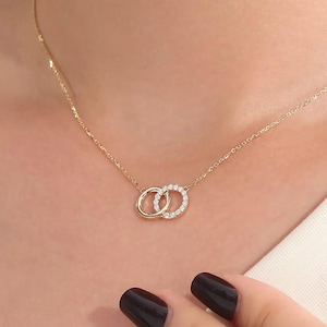 Puede incluir: Un collar de cadena dorada con dos colgantes circulares entrelazados. Un círculo es una banda de oro pulido liso, mientras que el otro es una banda de oro incrustada con pequeñas piedras claras y brillantes a lo largo de toda su circunferencia.
