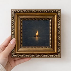 May include: A small square painting of a campfire burning in the dark, set within an ornate, antique-style gold frame with intricate scrollwork and beaded detailing along the inner edge, creating a contrast between the warm fire and dark background.