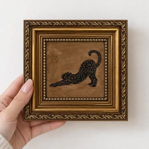 May include: A square painting in an ornate gold frame featuring a black leopard in a stretching pose against a solid brown background. The leopard is depicted in profile with its front paws extended forward and its tail curved upward.