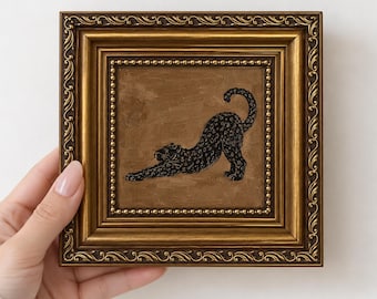 May include: A square painting in an ornate gold frame featuring a black leopard in a stretching pose against a solid brown background. The leopard is depicted in profile with its front paws extended forward and its tail curved upward.