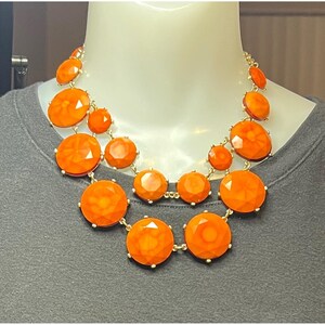 May include: An orange statement necklace with large, faceted, round and oval-shaped beads. The beads are connected by a gold-coloured chain. The necklace is displayed on a mannequin.