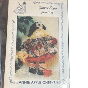 May include: A package featuring a handmade doll named Annie Apple Cheeks. The doll has blonde yarn hair, a black hat with a white flower, and a floral dress. It holds a small basket and a stuffed animal.