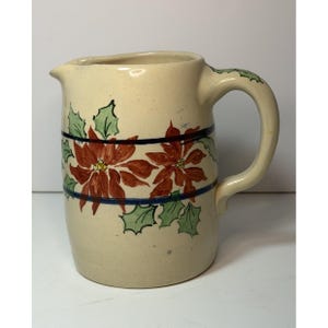 Puede incluir: Una jarra de cerámica de color crema con un diseño pintado a mano de flores de pascua rojas y hojas de acebo verdes. El patrón floral está bordeado por dos franjas horizontales de color azul oscuro que rodean el cuerpo de la jarra.