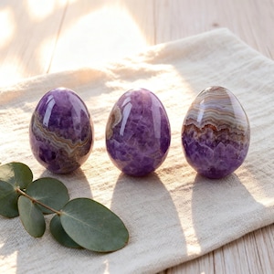 Puede incluir: Tres piedras pulidas en forma de huevo con capas de amatista púrpura profunda y ágata bandeada. Las piedras tienen una superficie lisa y reflectante con estructuras cristalinas visibles y variaciones de color naturales en tonos violeta, blanco y marrón.