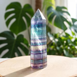 May include: A tall, faceted crystal tower with a pointed top. The crystal features bands of purple, blue, and green hues. It is displayed on a round, wooden surface, with green foliage in the background. The crystal is approximately 6 inches tall.