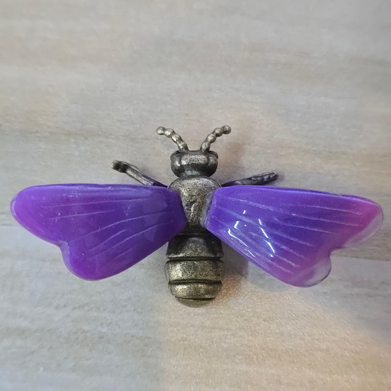 May include: A bee-shaped brooch featuring an antique bronze-toned metal body with segmented detailing and two large, translucent purple wings with etched linear patterns extending from the center of the insect's thorax.